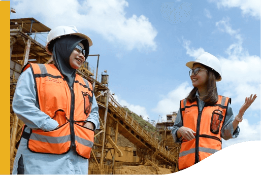 The image shows two individuals wearing orange safety vests and white hard hats standing in front of an industrial site with machinery and structures. The sky is partly cloudy.