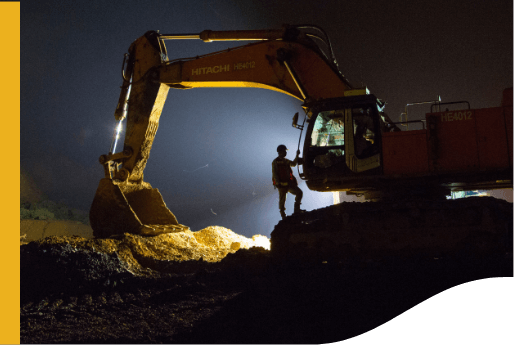 The image shows a construction site at night with an excavator in operation. The excavator, labeled "HITACHI" with model number "HE4012," has a person standing on it, silhouetted against a bright light source behind them. The excavator's bucket is filled with dirt or debris, which is illuminated by the same light.