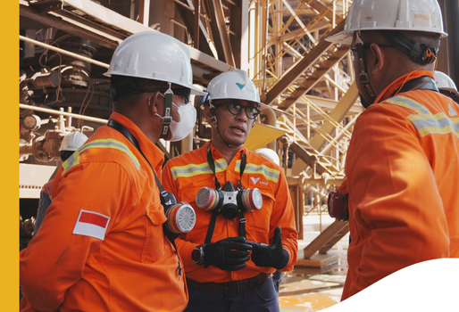 Three individuals wearing orange safety uniforms and white helmets are engaged in a discussion at an industrial site. The person in the center is wearing a face mask and has two respirators hanging from the neck. The individual on the left has an Indonesian flag patch on the sleeve.