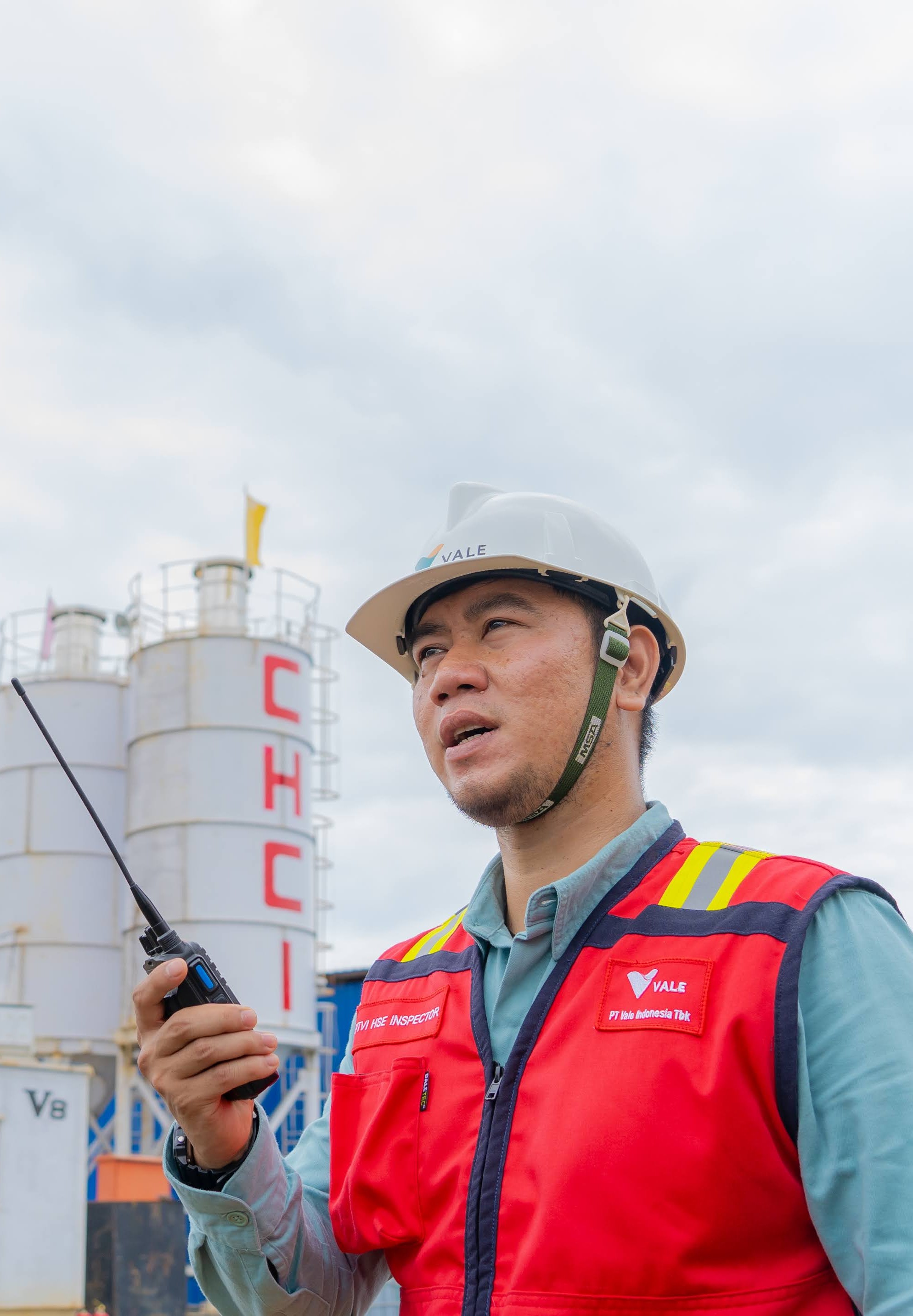 A PT Vale HSE inspector wearing a Vale hard hat and red safety vest holds a handheld radio in front of the CHCI plant facility.