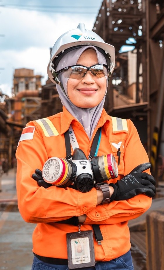 A female worker wearing a hijab, a Vale hard hat, safety glasses, and orange PPE holds a respirator mask and smiles at an industrial plant.